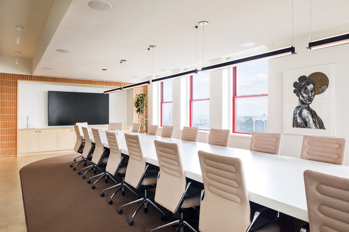 A long white conference table surrounded by white office chairs. Three windows overlook a city skyline.